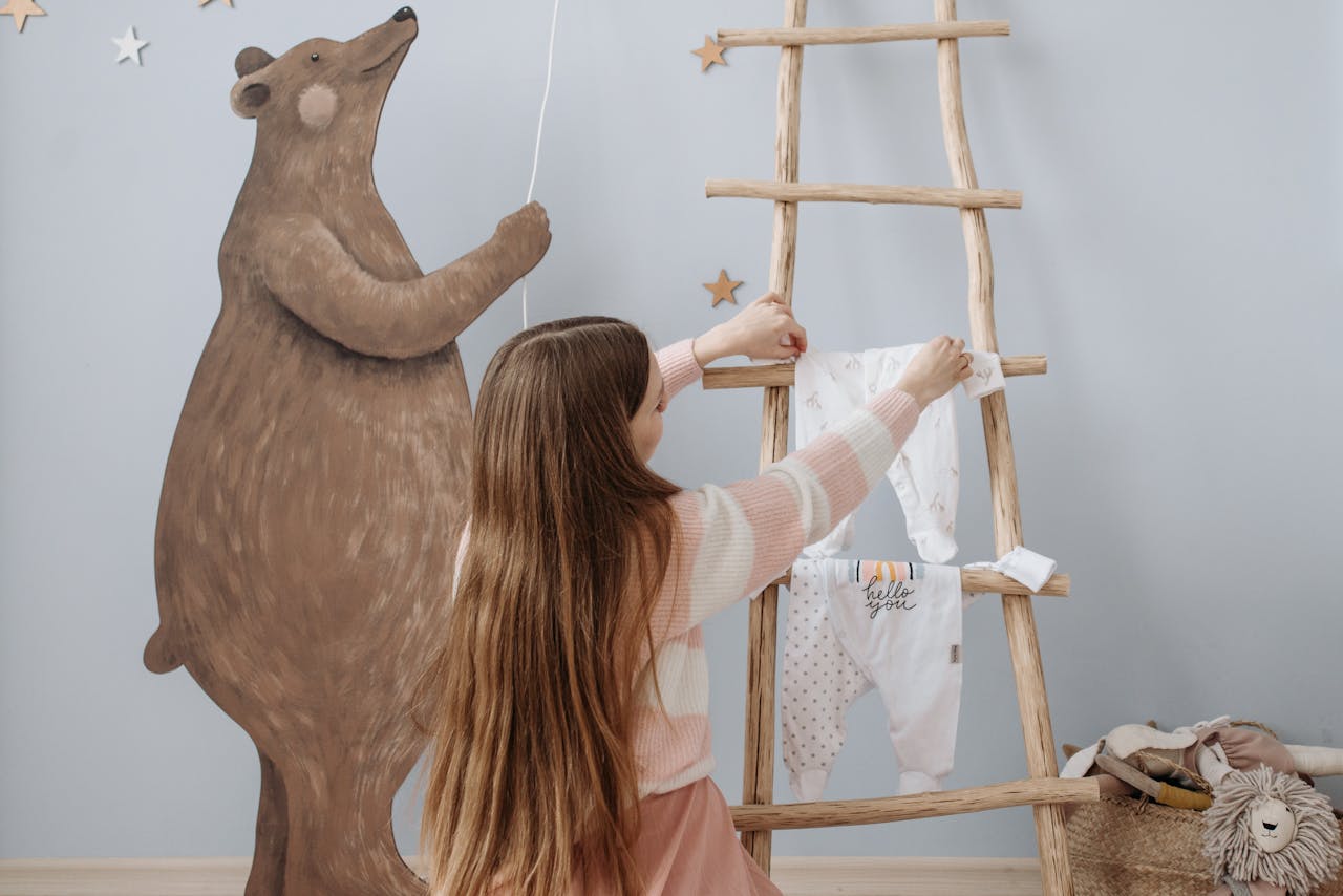 Woman organizes baby clothes on wooden ladder in decorated nursery, cozy and playful environment.